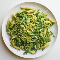 Spring Green Pesto Pasta Salad in a serving bowl, mixed with bright green peas, peppery arugula, and toasted pine nuts.